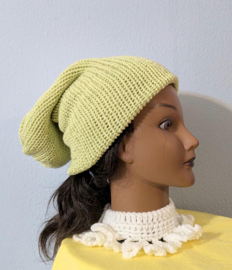Puede incluir: Un gorro tejido de color verde claro. El gorro est&aacute; hecho de una tela de punto acanalado y tiene un ajuste holgado. El gorro se muestra en una cabeza de maniqu&iacute; con cabello oscuro y un cuello de ganchillo blanco.