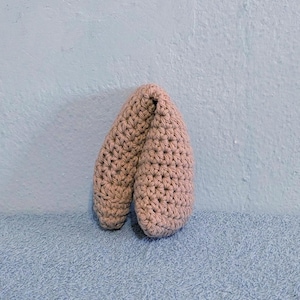 May include: A soft, light brown crochet object with a rounded, folded shape. The item features a dense, textured stitch pattern throughout its surface, creating a uniform appearance. The structure is curved and compact, showing consistent yarn work.