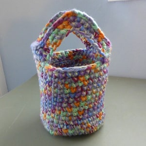 May include: A handmade crochet basket in a cylindrical shape with a handle. The basket is made of yarn in a variety of colors, including purple, orange, green, and blue. The basket is approximately 6 inches tall and 4 inches in diameter.