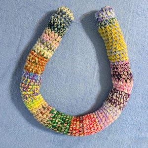 May include: A colorful, horseshoe-shaped crochet item. The item is made of yarn in various colors, including blue, yellow, pink, green, and orange. The crochet stitches are visible, creating a textured surface. The item is likely a decorative piece or a craft project.