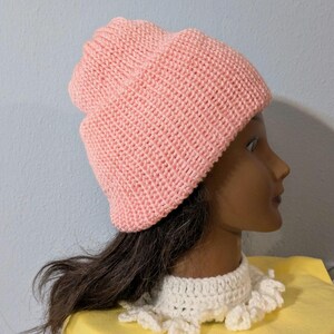 May include: A light pink knitted beanie hat. The hat is made of a textured knit fabric and has a folded brim. The hat is displayed on a mannequin head with dark hair and a white crocheted collar.