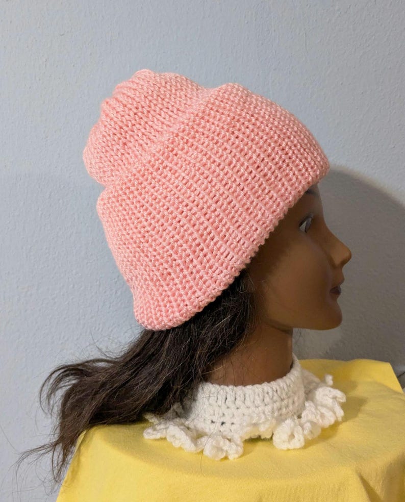 May include: A light pink knitted beanie hat. The hat is made of a textured knit fabric and has a folded brim. The hat is displayed on a mannequin head with dark hair and a white crocheted collar.