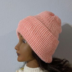 May include: A light pink knitted beanie hat. The hat is made of a soft, textured yarn and features a folded brim. The beanie is displayed on a mannequin head with dark hair and a white crocheted collar.