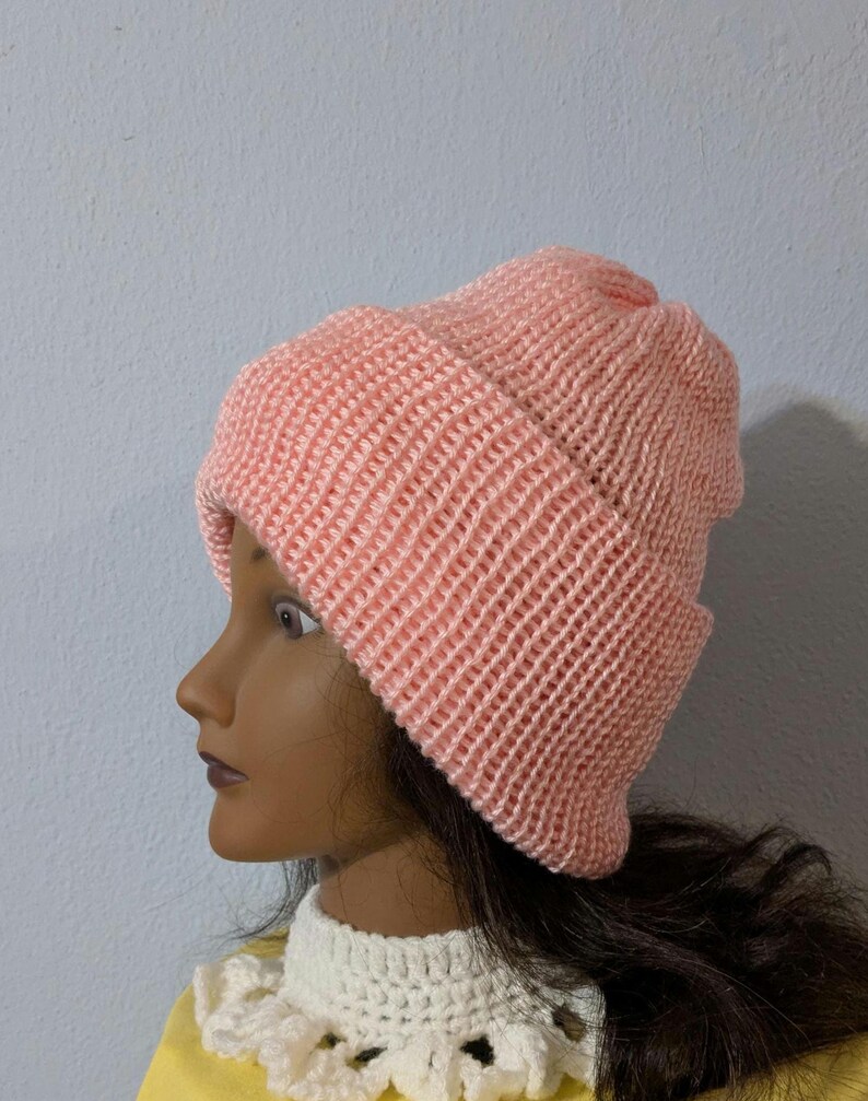 May include: A light pink knitted beanie hat. The hat is made of a soft, textured yarn and features a folded brim. The beanie is displayed on a mannequin head with dark hair and a white crocheted collar.