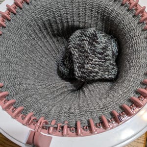 May include: A circular knitting loom with a partially completed gray and white knitted hat. The loom is white with pink pegs, and the hat is nestled inside the knitted fabric. The yarn is a mix of gray and white.