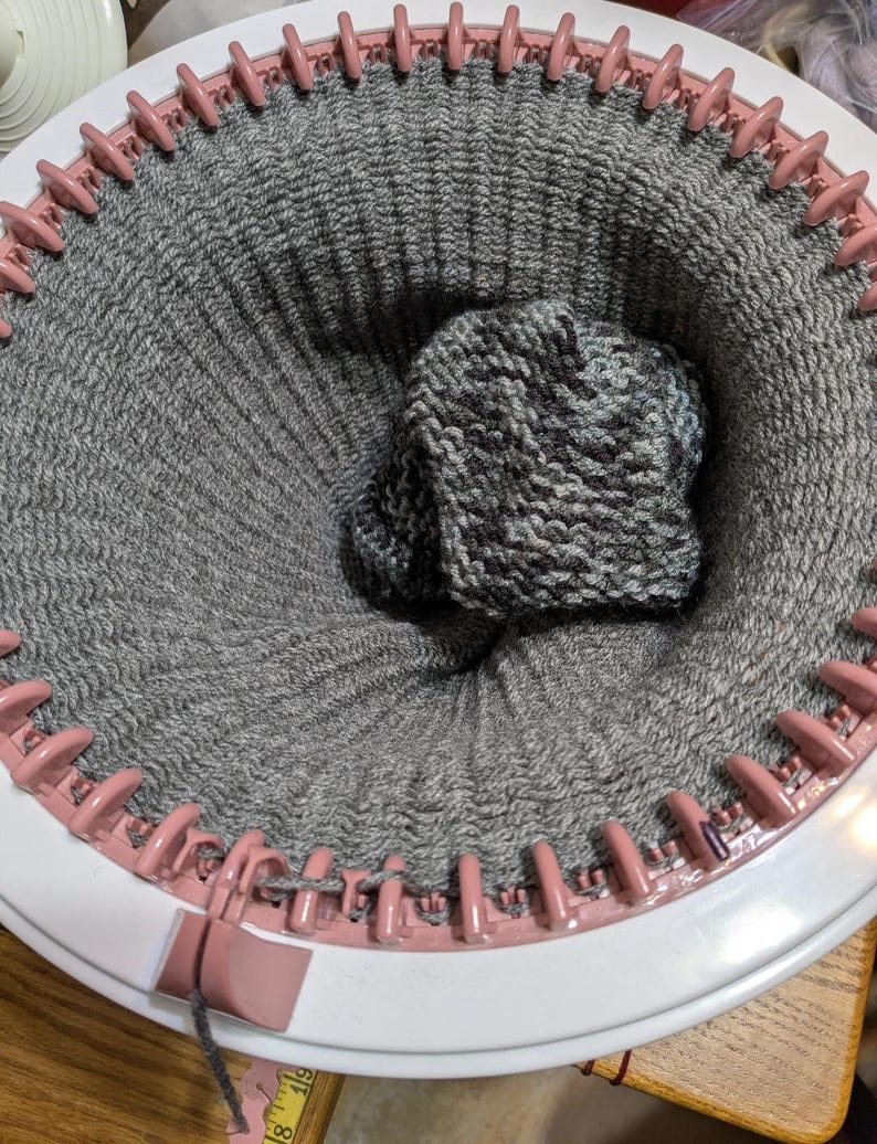 May include: A circular knitting loom with a partially completed gray and white knitted hat. The loom is white with pink pegs, and the hat is nestled inside the knitted fabric. The yarn is a mix of gray and white.