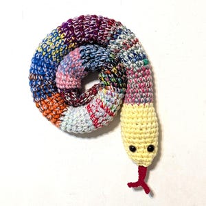 May include: A colorful, crocheted snake toy. The body is a spiral of various colors including blue, orange, pink, and purple. The head is yellow with black button eyes and a red forked tongue. The snake is a handmade craft item.