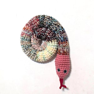 May include: A coiled crochet snake toy featuring a solid pink head with black plastic eyes and a red forked tongue. The body is crafted from variegated yarn in a mix of blue, brown, cream, and red tones, creating a textured, multi-colored pattern.