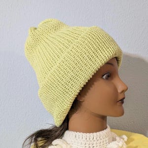 Puede incluir: Un gorro tejido de color verde claro con un borde doblado. El gorro est&aacute; hecho de una tela de punto texturizada y se muestra en una cabeza de maniqu&iacute;. El maniqu&iacute; lleva un cuello de ganchillo blanco y una camisa amarilla.