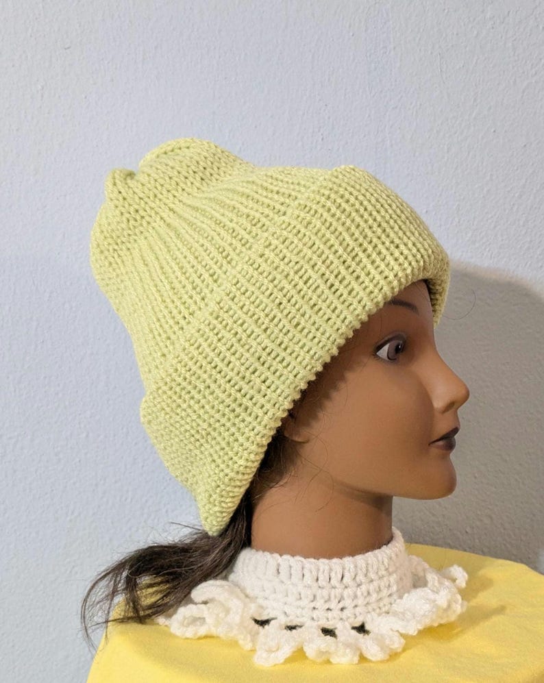 Puede incluir: Un gorro tejido de color verde claro con un borde doblado. El gorro est&aacute; hecho de una tela de punto texturizada y se muestra en una cabeza de maniqu&iacute;. El maniqu&iacute; lleva un cuello de ganchillo blanco y una camisa amarilla.