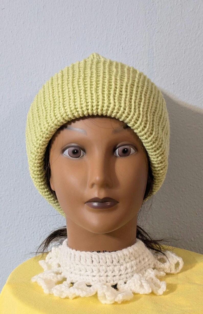 May include: A light green, ribbed knit beanie hat. The hat is worn by a mannequin with a cream-colored crocheted collar and a yellow shirt. The hat is a solid color and has a simple, classic design.
