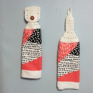May include: Two hanging kitchen towels with a white crochet top and a fabric bottom. The fabric features a geometric design with coral, black, and white patterns. One towel has a button closure.