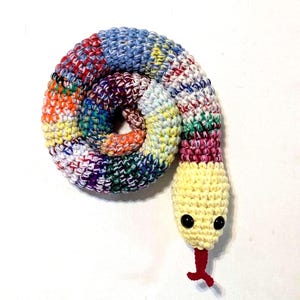 May include: A colorful, crocheted snake toy. The body is a mix of blue, red, orange, green, and yellow yarn, coiled on itself. The head is yellow with black button eyes and a red tongue. A handmade textile art piece.