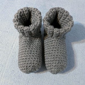 May include: A pair of gray, handmade crochet slippers. The slippers have a textured, ribbed appearance and a folded-over cuff at the top. The slippers are designed to keep feet warm and comfortable.