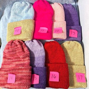 May include: A collection of handmade knit beanies in various colors, including light blue, pink, red, purple, and yellow. Each beanie has a folded brim and a small pink tag with a number on it. The beanies are arranged on a light blue surface.