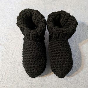 May include: A pair of black crochet slippers. The slippers are made of a thick, textured yarn and have a bootie-like design with a rolled cuff. The slippers are designed to keep feet warm and comfortable.
