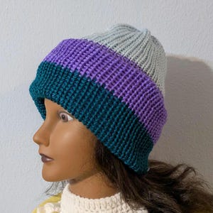 May include: A knitted beanie hat with a color block design. The hat features bands of teal, purple, and gray. The hat is made of a soft, textured yarn. This hat is a stylish accessory for cold weather.