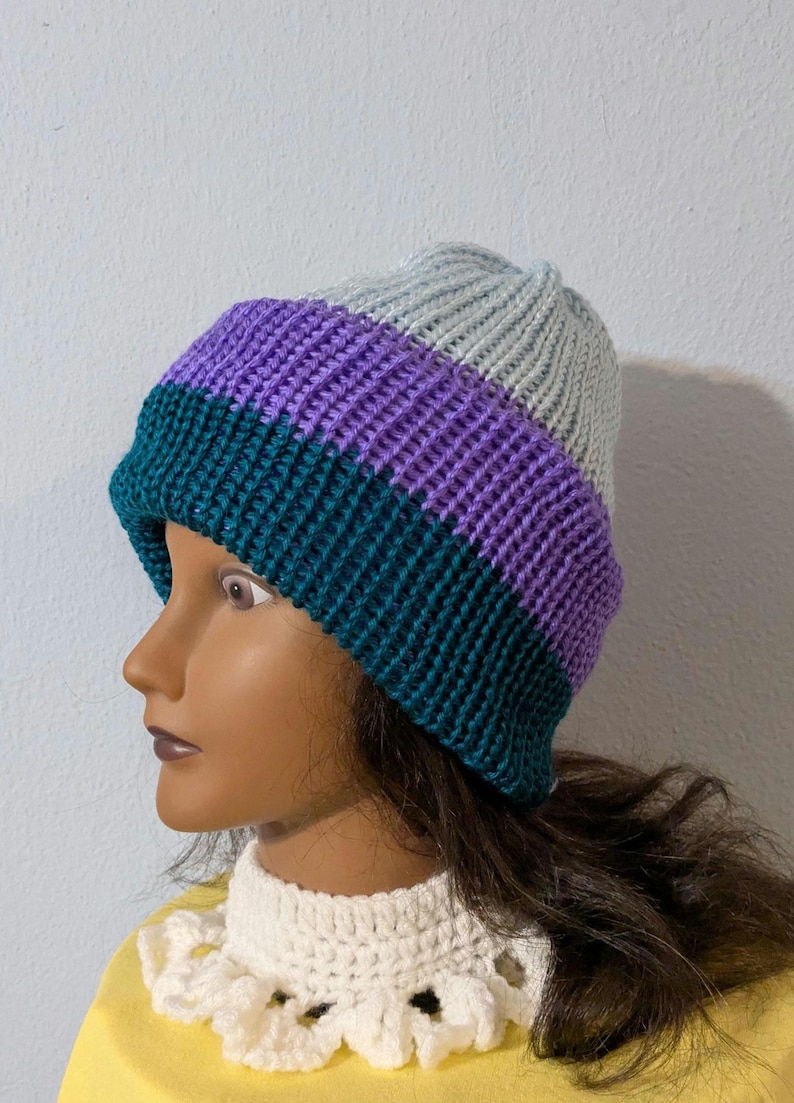 May include: A knitted beanie hat with a color block design. The hat features bands of teal, purple, and gray. The hat is made of a soft, textured yarn. This hat is a stylish accessory for cold weather.