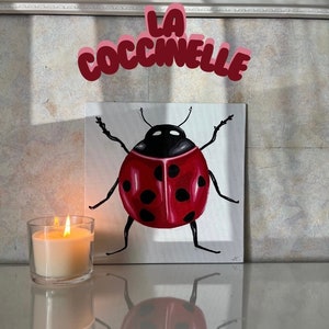 Op de afbeelding: Een schilderij van een rode lieveheersbeestje met zwarte stippen op een wit doek. Het schilderij is getiteld "La Coccinelle" in roze en rode letters.