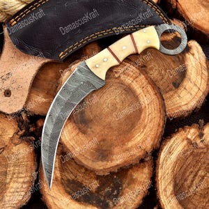 May include: A curved karambit knife with a patterned blade and bone handle, resting on a pile of wood slices. The knife has a finger ring and is accompanied by a leather sheath. The text "DamascusKraft" is visible.