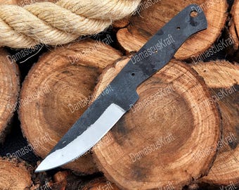 May include: A black, unfinished knife blade with a silver edge, resting on a bed of wood slices. A thick, beige rope is in the background. The knife has a hole near the handle and the text "DamascusKraft" is visible.