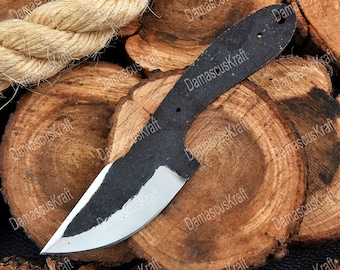 May include: A black, unfinished knife blade with a silver edge, resting on a wooden surface. The knife has a curved handle and a pointed tip. The blade is made of metal. The background includes a rope and wood slices. The text "DamascusKraft" is visible.