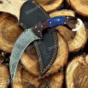May include: A Damascus steel karambit knife with a curved blade and a blue and brown handle. The knife rests in a black leather sheath with yellow stitching, set against a wood background. The text "DamascusKraft" is visible.