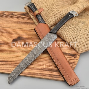 May include: A Damascus steel bread knife with a serrated blade and a dark wooden handle. The knife is in a brown leather sheath, resting on a wooden cutting board. The text "DAMASCUS KRAFT" is visible on the board.