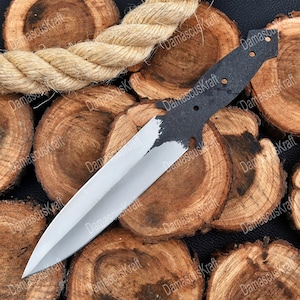 May include: A partially finished knife blade with a silver edge and a dark, textured handle. The knife rests on a bed of wood slices, with a thick rope in the background. The blade is sharp and pointed.