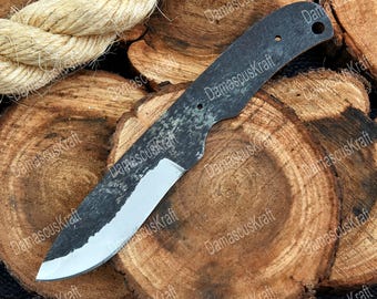 May include: A dark, unfinished knife blade with a textured, blackened surface and a polished, silver edge. The blade is set against a backdrop of wooden cross-sections and a thick rope.