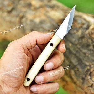 May include: A handcrafted knife with a cream-colored handle and a silver blade. The handle has two dark holes. The text "Damascus Kraft" is visible in the image. The knife is held in a hand.