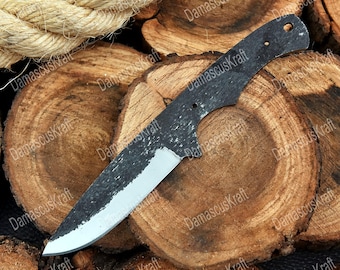 May include: A black and silver knife blade with a textured finish. The blade is set against a backdrop of wood slices and rope. The knife has a sharp edge and a handle with pre-drilled holes.