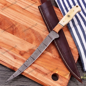 May include: A Damascus steel hunting knife with a wooden handle and a white bone pommel. The knife is on a wooden cutting board with a brown leather sheath.