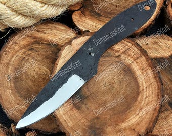 May include: A black, unfinished knife blade with a white edge, resting on a wooden surface. The blade has a hole near the handle and is made of dark metal. A length of rope is visible in the background.