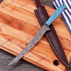 May include: A blue and silver Damascus steel knife with a turquoise handle and a leather sheath. The knife is on a wooden cutting board.