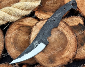 May include: A Damascus Kraft knife blade with a curved edge and a partially sharpened, bright silver blade. The dark, textured steel blade has three holes and rests on a bed of wood slices. A thick rope is in the background.