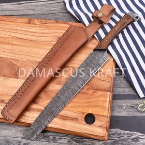 May include: A Damascus Kraft chef's knife with a patterned blade and a wooden handle, resting on a wooden cutting board. The knife is accompanied by a brown leather sheath. The text "DAMASCUS KRAFT" is visible on the cutting board.