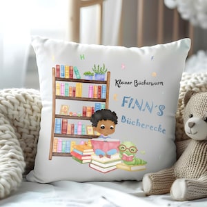 以下が含まれることがあります： 本棚、本を読んでいる子供、そして「Kleiner Bücherwurm FINN'S Bücherecke」というテキストが描かれたカラフルなイラストが施された白い枕です。
