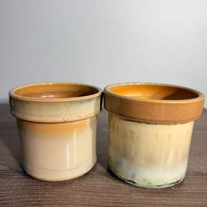 May include: Two cylindrical ceramic pots with thick rims. The left pot features a smooth gradient of cream and orange-brown glaze. The right pot displays a textured, mottled finish transitioning from a solid brown rim to a cream and pale green base.