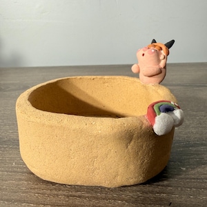 Puede incluir: Un pequeño cuenco redondo de cerámica de color beige con una superficie texturizada. Una pequeña figura rosa con cuernos negros y pelo naranja está en el borde. Una decoración en forma de arcoíris está en el lateral. El cuenco está sobre una superficie de madera.