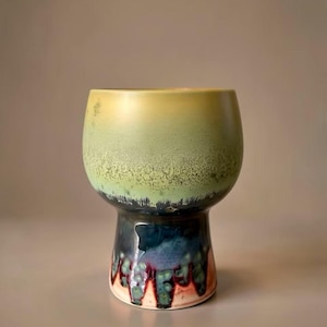 May include: A ceramic goblet with a rounded bowl and a flared base. The upper bowl features a gradient from yellow to light green with a textured finish. The lower base displays a dark blue glaze with vertical drips of green and reddish-brown.
