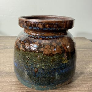 May include: A handcrafted ceramic jar with a wide, rounded body and a slightly flared rim. The jar features a rich, earthy color palette, with shades of brown, green, and blue. The glaze has a mottled, textured appearance, adding to its rustic charm.