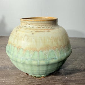 May include: A round ceramic vase featuring a wide, bulbous body with a short, narrow neck. The glaze transitions from a creamy, light beige at the top to a soft, muted sage green at the base, with visible drip patterns running down the sides.