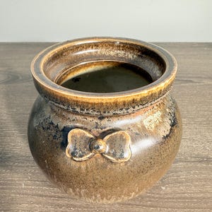 May include: A round ceramic pot with a wide opening and a thick, rolled rim. The exterior features a mottled brown and black glaze with a glossy finish. A small, molded bow-shaped detail is centered on the front of the pot's rounded body.