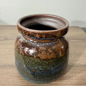 May include: A handcrafted ceramic jar with a wide opening and a rounded body. The jar features a textured surface with a blend of brown, green, and blue hues. The rim is a darker shade of brown. This decorative piece is suitable for various uses.