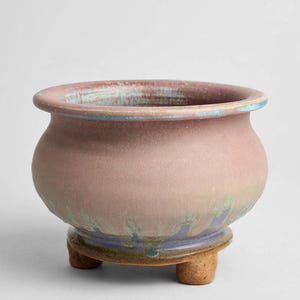 May include: A rounded ceramic planter with a wide rim, featuring a matte dusty pink finish that transitions into a blue and green drip glaze near the base. The vessel is supported by three short, sturdy, unglazed tan clay feet.