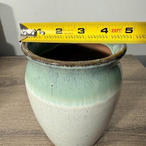 May include: A ceramic vase with a two-tone design. The top half is a light seafoam green, transitioning to a creamy white base. The rim is a dark brown. The vase is approximately 4 inches in diameter.