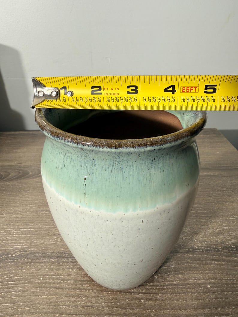 May include: A ceramic vase with a two-tone design. The top half is a light seafoam green, transitioning to a creamy white base. The rim is a dark brown. The vase is approximately 4 inches in diameter.