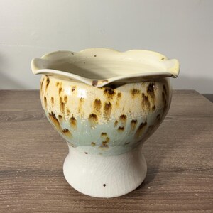 May include: A ceramic bowl with a scalloped rim. The bowl features a cream-colored interior and a speckled exterior with shades of tan, brown, and a touch of light blue. The base is white and slightly cracked.