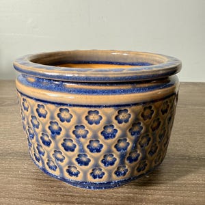 May include: A round ceramic planter featuring a two-tone glaze with a tan base and a deep blue finish on the raised rim and embossed flower patterns. The interior of the pot is glazed in a solid tan color, and the exterior displays a repeating floral motif.